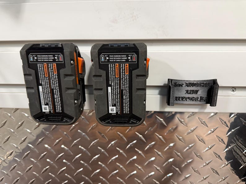 Puede incluir: Dos bater&iacute;as de herramientas el&eacute;ctricas negras y grises con detalles en naranja est&aacute;n montadas en una pared, junto a un soporte de bater&iacute;a negro impreso en 3D. Las bater&iacute;as tienen etiquetas de advertencia e informaci&oacute;n del producto. El soporte tiene texto.