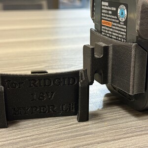 Puede incluir: Soporte de bater&iacute;a negro impreso en 3D para bater&iacute;as Ridgid 18V Hyper Lithium. El soporte est&aacute; dise&ntilde;ado para sujetar la bater&iacute;a de forma segura. El texto "For RIDGID 18V HYPER Li" est&aacute; impreso en el soporte.