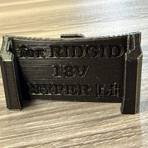 Puede incluir: Soporte negro impreso en 3D para bater&iacute;as RIDGID 18V HYPER Li. El soporte tiene una parte superior curvada y una forma rectangular con el texto "for RIDGID 18V HYPER Li" en relieve en la parte delantera. Est&aacute; dise&ntilde;ado para sujetar una bater&iacute;a.
