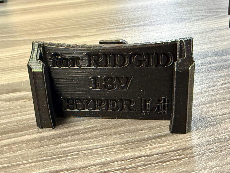 Puede incluir: Soporte negro impreso en 3D para bater&iacute;as RIDGID 18V HYPER Li. El soporte tiene una parte superior curvada y una forma rectangular con el texto "for RIDGID 18V HYPER Li" en relieve en la parte delantera. Est&aacute; dise&ntilde;ado para sujetar una bater&iacute;a.