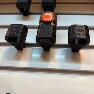 May include: Black 3D-printed accessories for a RIGID 12V HYPER LITE tool. The image shows three black components, including two caps and a connector with an orange button. The accessories are mounted on a white surface.