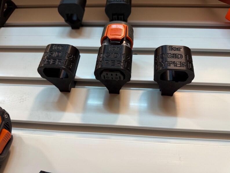 May include: Black 3D-printed accessories for a RIGID 12V HYPER LITE tool. The image shows three black components, including two caps and a connector with an orange button. The accessories are mounted on a white surface.