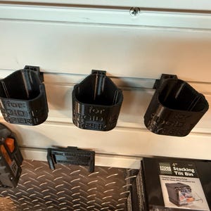May include: Three black 3D-printed tool holders mounted on a white wall. Each holder is labeled with text. A battery pack and a stacking tilt bin are also visible.