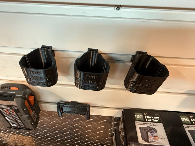 May include: Three black 3D-printed tool holders mounted on a white wall. Each holder is labeled with text. A battery pack and a stacking tilt bin are also visible.