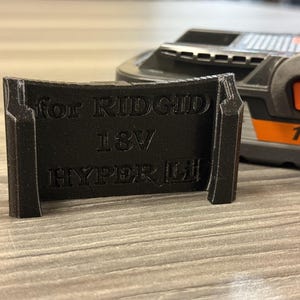 Puede incluir: Un adaptador negro impreso en 3D para bater&iacute;as RIDGID 18V HYPER Li. El adaptador es rectangular con el texto "for RIDGID 18V HYPER Li". Una herramienta el&eacute;ctrica naranja y negra es parcialmente visible en el fondo.