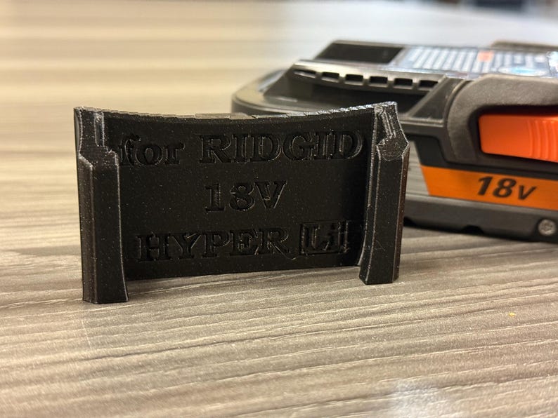 Puede incluir: Un adaptador negro impreso en 3D para bater&iacute;as RIDGID 18V HYPER Li. El adaptador es rectangular con el texto "for RIDGID 18V HYPER Li". Una herramienta el&eacute;ctrica naranja y negra es parcialmente visible en el fondo.