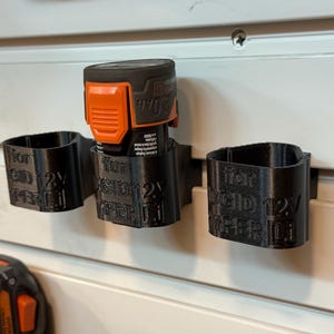 May include: Black 3D-printed battery holders for a power tool, mounted on a white wall. The holders are labeled "for RIDGID 12V HYPER Li". An orange and black battery is inserted into one of the holders.