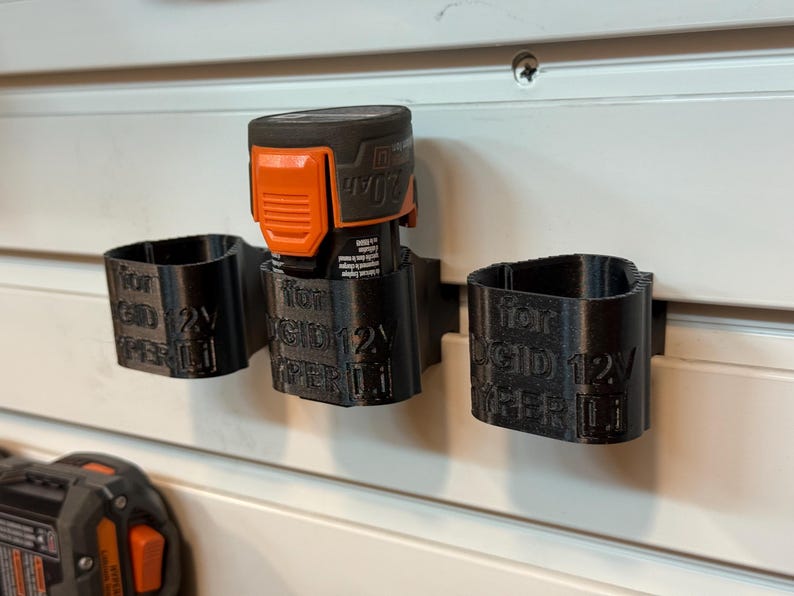 May include: Black 3D-printed battery holders for a power tool, mounted on a white wall. The holders are labeled "for RIDGID 12V HYPER Li". An orange and black battery is inserted into one of the holders.