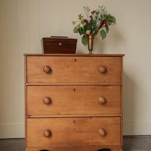 May include: A light brown wooden chest of drawers featuring three horizontal drawers, each equipped with two round wooden knobs and a central keyhole. A dark brown wooden box and a brass vase containing a floral arrangement sit on top.