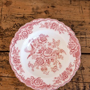 May include: White ceramic plate with a red floral and bird pattern. The plate has a scalloped edge and a detailed design featuring flowers and birds. The plate is set on a wooden surface.