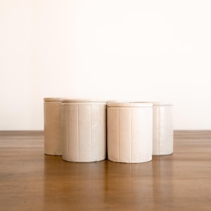 May include: Four off-white, cylindrical stoneware crocks with lids, arranged on a wooden surface. The crocks have vertical ribbed detailing and a rustic, vintage aesthetic. The background is a plain white wall.