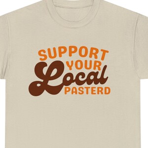 May include: A beige t-shirt with the text "Support Your Local" in brown and orange lettering. The word "Local" is in a retro, stylized font.