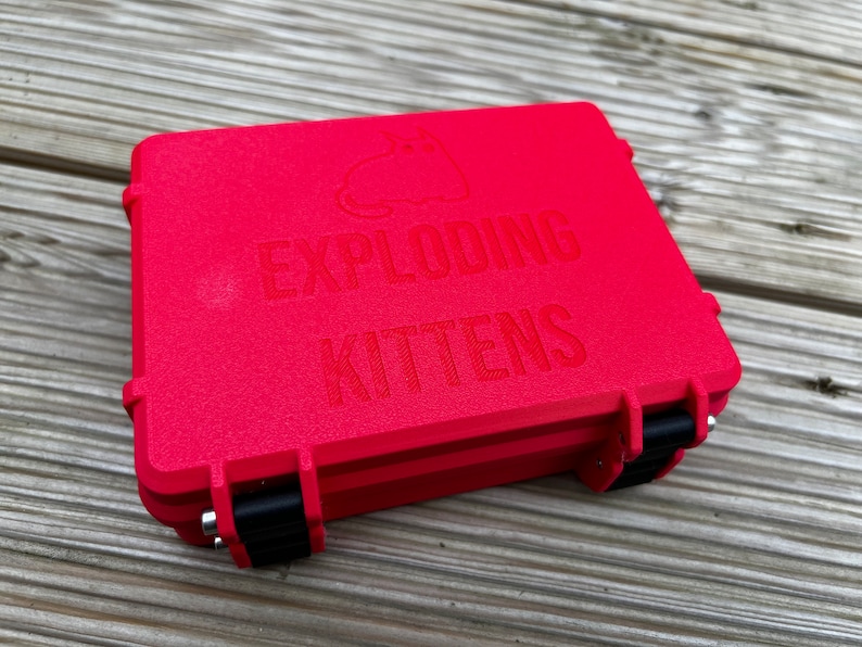 May include: A red plastic case with black latches. The case has the words "Exploding Kittens" embossed on the top.