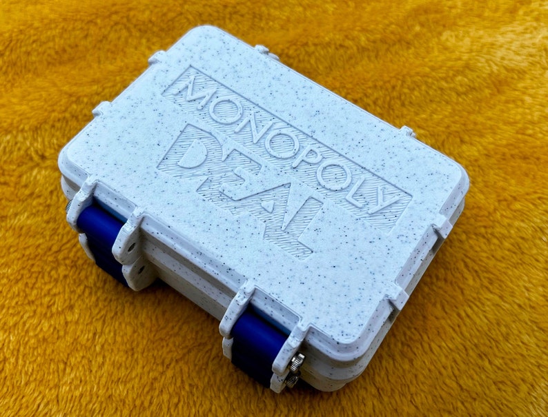 Op de afbeelding: Een witte Monopoly Deal-speldoos met een gespikkelde textuur en blauwe scharnieren. Op de doos staat "MONOPOLY DEAL" gedrukt.