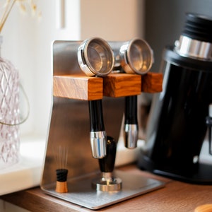 Könnte beinhalten: Ein Edelstahl-Espresso-Portafilterhalter mit zwei Portafiltern und einem Holzsockel. Der Halter steht auf einer Holztheke.