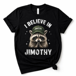 May include: A black t-shirt featuring a graphic of a raccoon wearing a green baseball cap with the text JIMOTHY on it. Above the raccoon, the text reads I BELIEVE IN, and below the raccoon, the text reads JIMOTHY. Small white stars surround the raccoon.