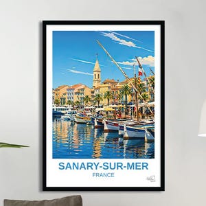 Op de afbeelding: Een ingelijste poster met een levendige illustratie van Sanary-sur-Mer, Frankrijk. Het kunstwerk toont een haven met boten, gebouwen en een kerktoren onder een helderblauwe lucht. De poster bevat de tekst "SANARY-SUR-MER FRANCE".