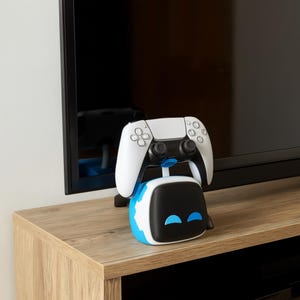 Pode incluir: Um controle de videogame branco e preto repousa sobre um suporte de carregamento azul, preto e branco. O suporte tem um formato arredondado com detalhes em azul e está colocado sobre uma superfície de madeira marrom clara. O fundo inclui uma tela de televisão preta.