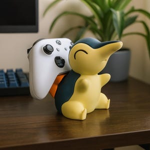 May include: A yellow and navy blue Cyndaquil-shaped controller holder with an orange accent, holding a white Xbox controller. The holder is on a dark brown wooden surface.