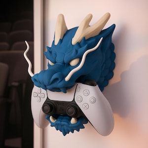 May include: A blue dragon head wall mount holding a white and black game controller. The dragon has cream-coloured horns and whiskers. The controller has a black centre with white buttons and directional pads. The dragon is mounted on a white wall.