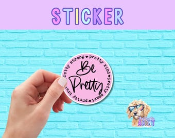 May include: A round pink sticker with the words "Be Pretty" in a cursive font, surrounded by the words "pretty, strong, kind, smart, brave." The word "STICKER" is at the top. A cartoon dog wearing sunglasses is in the lower right corner.