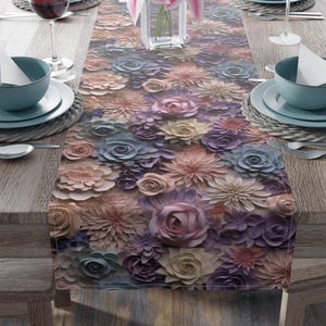 May include: A floral table runner with a 3D effect, featuring a variety of pastel-colored flowers in shades of pink, purple, and beige. The runner is placed on a wooden table set for a meal, with plates, bowls, and wine glasses.