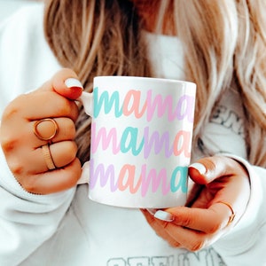 May include: White ceramic mug with the word "mama" repeated in pastel colors: teal, pink, and lavender. The mug is held by a person wearing a white sweatshirt. The mug is a standard size for coffee or tea.