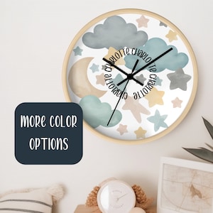May include: A white clock with a wooden frame features a watercolor design of clouds, stars, and a crescent moon. The clock face is surrounded by the name "Charlotte" written in a circular pattern.