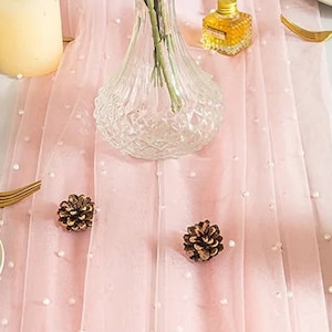 May include: A pink table runner with white pearl accents, two white candles, and a clear glass vase with white flowers. The runner is on a white tablecloth with gold silverware and two white plates.
