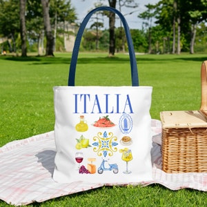 K&ouml;nnte beinhalten: Wei&szlig;e Tragetasche mit blauen Griffen mit dem Wort "ITALIA" in Blau. Die Tasche ist mit Aquarellillustrationen italienischer Speisen, Sehensw&uuml;rdigkeiten und eines Rollers verziert. Die Tasche liegt auf einer Picknickdecke auf dem Gras.