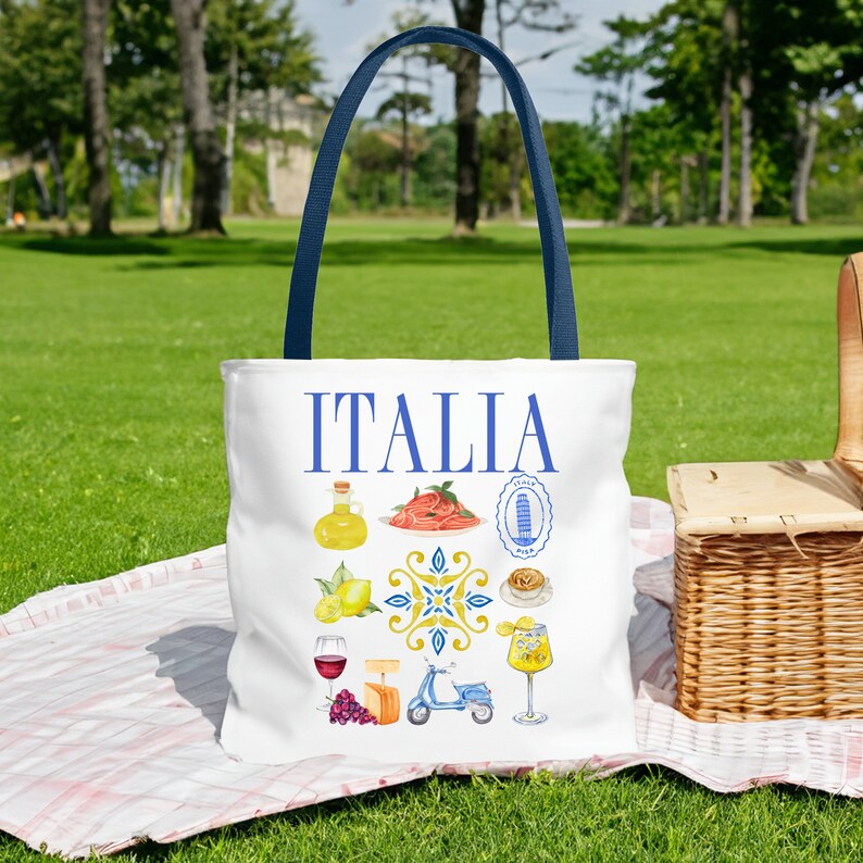 K&ouml;nnte beinhalten: Wei&szlig;e Tragetasche mit blauen Griffen mit dem Wort "ITALIA" in Blau. Die Tasche ist mit Aquarellillustrationen italienischer Speisen, Sehensw&uuml;rdigkeiten und eines Rollers verziert. Die Tasche liegt auf einer Picknickdecke auf dem Gras.