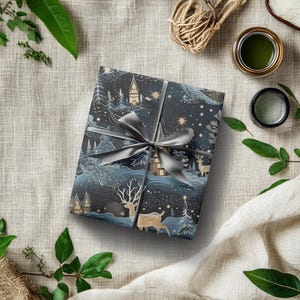May include: A wrapped gift with a dark blue and white winter scene design, tied with a gray ribbon. The wrapping paper features illustrations of houses, trees, deer, and stars. The gift is surrounded by greenery and a jar of green paint.