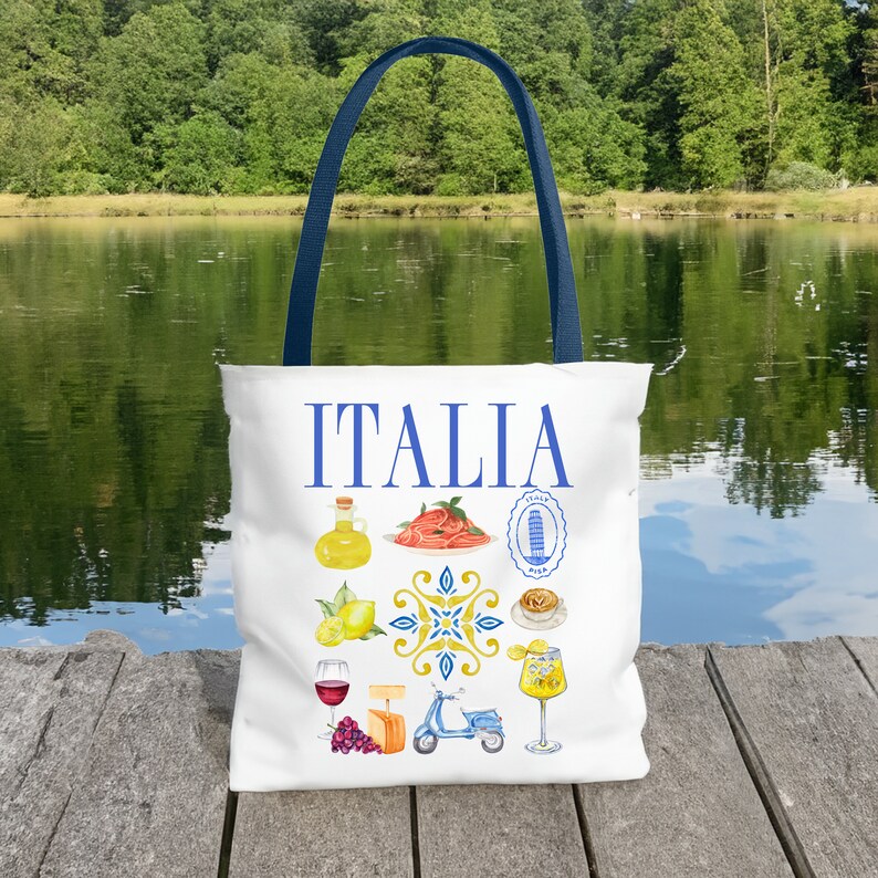 K&ouml;nnte beinhalten: Wei&szlig;e Tragetasche mit blauen Henkeln. Das Wort "ITALIA" ist blau auf der Tasche aufgedruckt. Sie zeigt Aquarellillustrationen italienischer Speisen und Sehensw&uuml;rdigkeiten, darunter Pasta, Zitronen, Wein und einen Roller. Die Tasche liegt auf einem Holzsteg.