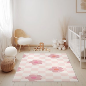 K&ouml;nnte beinhalten: Eine rosa-wei&szlig;e karierte Spielmatte mit rosa Blumenmustern. Die Matte liegt auf einem hellen Holzboden in einem Kinderzimmer mit einem wei&szlig;en Babybett, einem Korbstuhl und einer Holzspielzeugbahn.