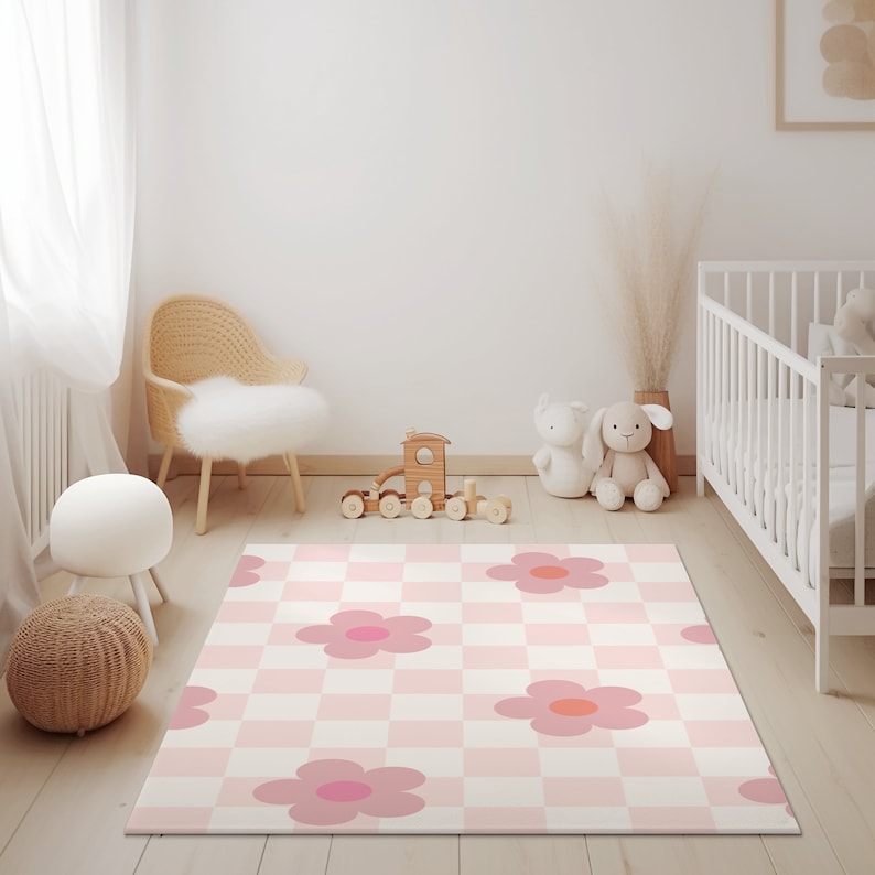 K&ouml;nnte beinhalten: Eine rosa-wei&szlig;e karierte Spielmatte mit rosa Blumenmustern. Die Matte liegt auf einem hellen Holzboden in einem Kinderzimmer mit einem wei&szlig;en Babybett, einem Korbstuhl und einer Holzspielzeugbahn.
