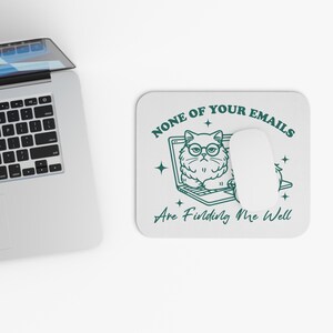 Op de afbeelding: Een witte muismat met een groene omtrek van een kat met een bril die op een laptop zit. De tekst "NONE OF YOUR EMAILS ARE FINDING ME WELL" is in het groen geschreven.
