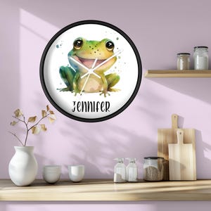 May include: A round wall clock with a black frame. The clock face features a watercolor illustration of a smiling green frog with the name "JENNIFER" in black script. The clock hands are white.