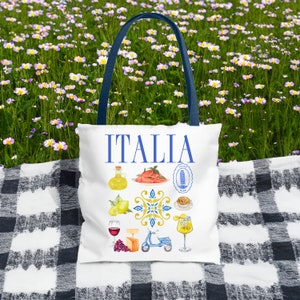K&ouml;nnte beinhalten: Wei&szlig;e Tragetasche mit blauen Griffen mit dem Wort "ITALIA" in Blau. Die Tasche ist mit Aquarellillustrationen von italienischem Essen, Sehensw&uuml;rdigkeiten und einem Roller verziert. Die Tasche liegt auf einer schwarz-wei&szlig; karierten Decke.