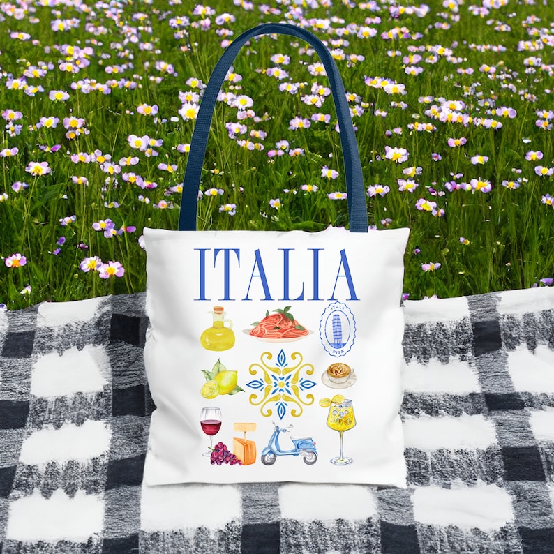 K&ouml;nnte beinhalten: Wei&szlig;e Tragetasche mit blauen Griffen mit dem Wort "ITALIA" in Blau. Die Tasche ist mit Aquarellillustrationen von italienischem Essen, Sehensw&uuml;rdigkeiten und einem Roller verziert. Die Tasche liegt auf einer schwarz-wei&szlig; karierten Decke.