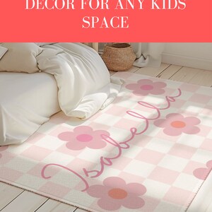 K&ouml;nnte beinhalten: Ein rosa-wei&szlig; karierter Teppich mit einem rosa Blumendesign und dem Namen "Isabella" in Kursivschrift. Der Teppich befindet sich in einem Schlafzimmer mit einem wei&szlig;en Bett und einem Weidenkorb.