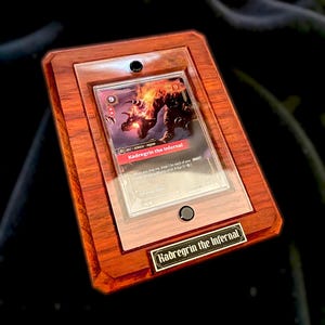 Pode incluir: Uma moldura de exibição retangular de madeira com uma capa protetora transparente contendo uma carta colecionável Kadregrin the Infernal. A moldura tem um acabamento polido e uma pequena placa de metal na parte inferior com o texto Kadregrin the Infernal.