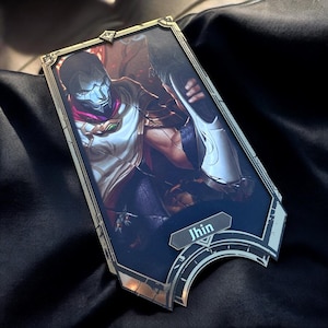 Puede incluir: Una tarjeta con marco dorado y fondo oscuro que presenta un personaje del videojuego League of Legends. El personaje lleva una máscara blanca y negra y una túnica blanca. La tarjeta tiene la palabra "Jhin" impresa.