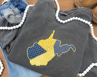 Könnte beinhalten: Ein dunkelgraues Sweatshirt mit einer gestickten Karte von West Virginia in gelbem und blauem Stoff mit Blumenmuster. Das Sweatshirt ist mit einer Jeans, einem grauen Scrunchie und einer weißen Perlenkette gefaltet.