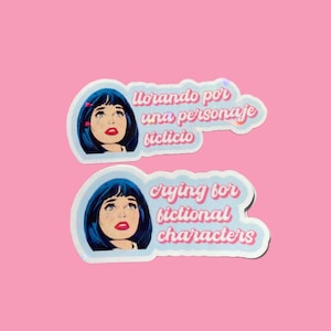 Op de afbeelding: Twee stickers met een pop-art stijl illustratie van een vrouw met blauw haar en tranen. De bovenste sticker leest "Llorando por una personaje ficticio" en de onderste sticker leest "crying for fictional characters". De achtergrond is roze.