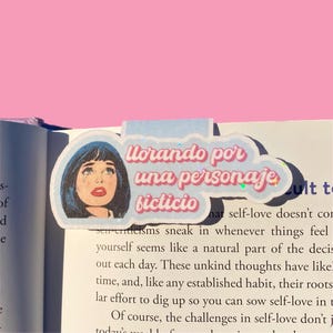 Op de afbeelding: Een sticker met een pop-art illustratie van een vrouw met blauw haar en tranen, met de Spaanse tekst "Llorando por una personaje ficticio" in roze en blauw. De sticker is op een boek geplaatst.