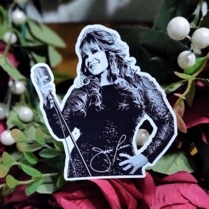 May include: Black and white sticker of a woman singing into a microphone. She is wearing a sparkly dress and has long hair. Her signature is visible in the bottom right corner.
