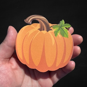 May include: A die-cut sticker shaped like a round orange pumpkin with a brown stem and a single green leaf attached to the top right side. The pumpkin features vertical ridges and a smooth, matte-like finish, presented against a black background.