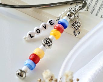 Könnte beinhalten: Ein Lesezeichen mit dem Wort "LOVE" aus weißen Buchstabenperlen. Das Lesezeichen hat bunte Perlen in Rot, Gelb und Blau sowie einen silbernen Blumenanhänger. Der Metall-Lesezeichenclip ist sichtbar.