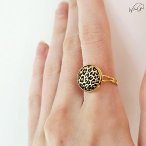 Könnte beinhalten: Ein goldfarbener Ring mit Leopardenmuster. Der Ring hat ein Doppelband und eine kreisförmige Fassung. Der Leopardenprint ist schwarz und beige. Der Ring ist ein Modeaccessoire.