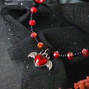 Könnte beinhalten: Ein schwarzes Perlenarmband mit roten facettierten Perlen und einem Herzanhänger mit silbernen Flügeln. Auf dem Armband steht der Text "Angel Devil Heart Bracelet Dark Romance Vibes WakaGirl".